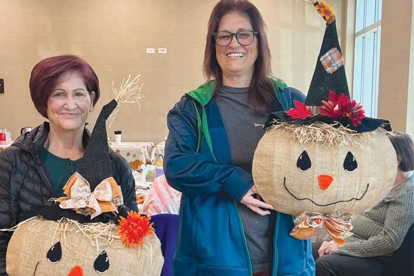 Two women smile while holding large burlap scarecrow crafts